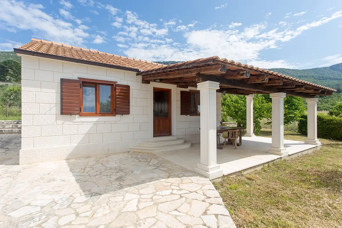Front view of the house with a stone facade, a column, outdoor dining table, entrance to the villa, and windows.
